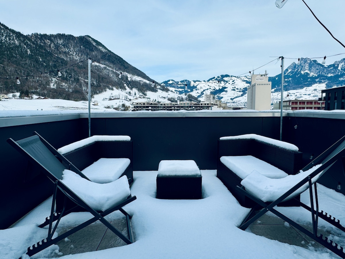 A balcony features two lounge chairs and a small table, all covered with a layer of fresh snow. The surrounding landscape showcases snow-capped mountains and a serene winter scene, providing a tranquil outdoor space for relaxation.