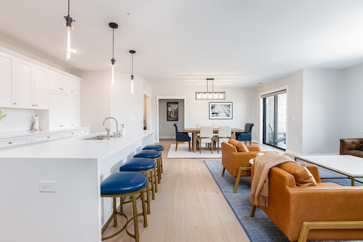 An open-concept living space showcases a modern kitchen with white cabinets and a long island. Four blue stools are positioned at the island. A dining area features a large table surrounded by six chairs, with a cozy seating area visible in the background.