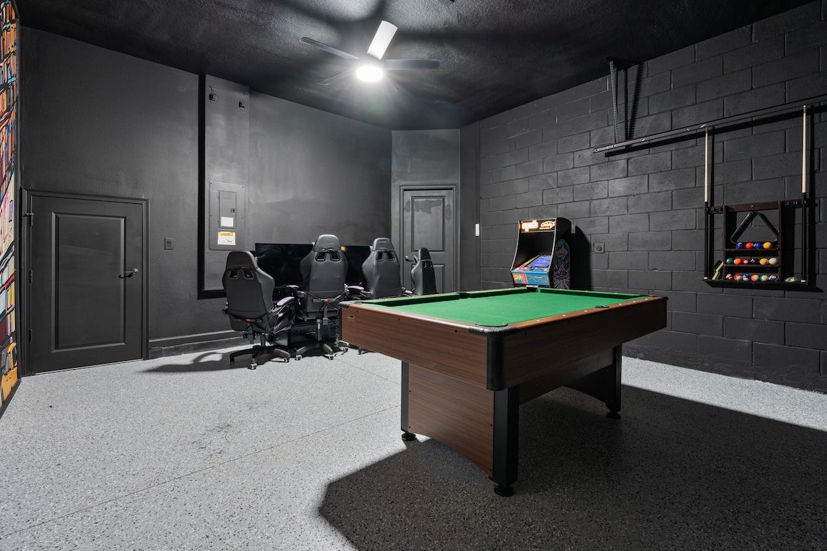 A spacious game room is featured, equipped with a green felt pool table at the center. Black walls provide a modern backdrop, while seating is arranged around a large TV. An arcade game machine and a rack for billiard balls are visible, enhancing the entertainment options.