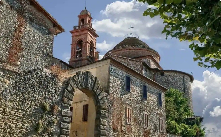 An ancient stone building is depicted, showcasing a mixture of architectural styles. A bell tower rises prominently, while a rounded dome is visible nearby. Lush greenery lines the structure, and an archway is present in the foreground, adding depth to the imagery.