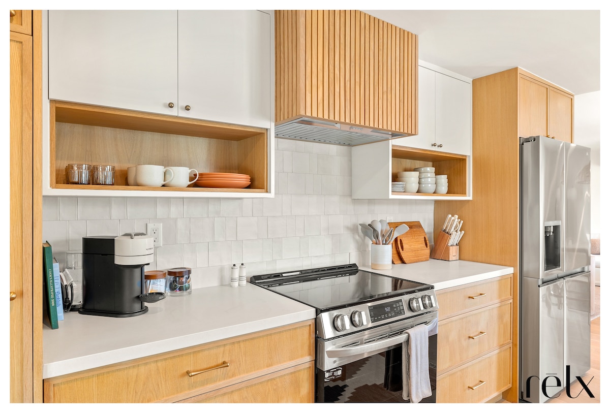 A modern kitchen features custom white oak cabinetry contrasted by light countertops. Open shelving displays dishware and accessories. A stainless steel refrigerator stands beside a sleek gas range with a hood above. A coffee maker is positioned nearby, enhancing the functional design.