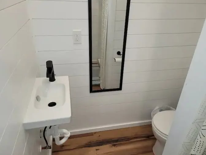 A compact bathroom is featured, with a modern sink and a large mirror mounted on the wall. Light-colored wood flooring contrasts with the white shiplap walls. A shower can be seen through a curtain, while a wastebasket is positioned discreetly nearby.
