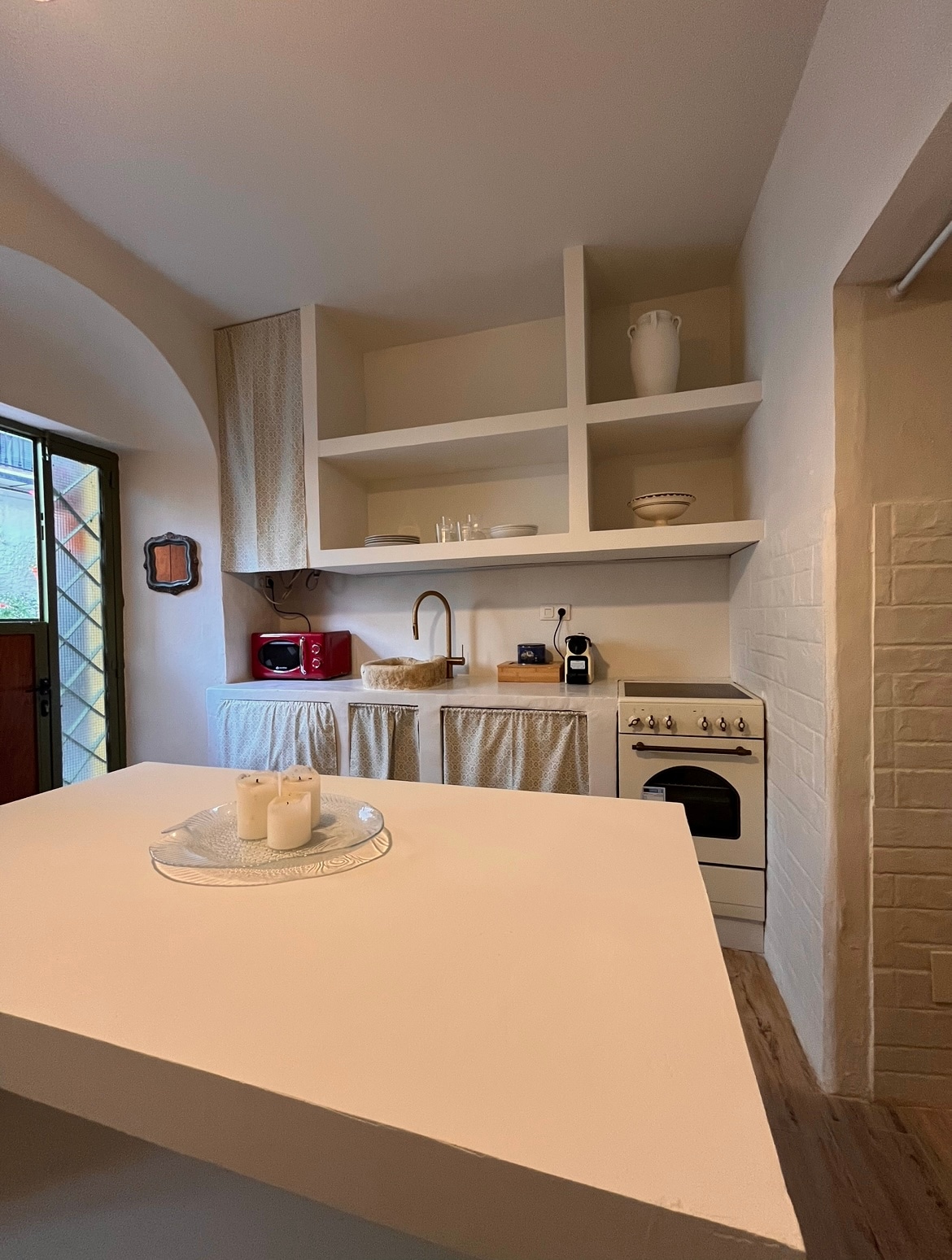 A cozy kitchen area features light-colored cabinetry with open shelving. A small sink and an electric stove are included, along with a coffee maker. A decorative tray with candles is placed on a central island, while natural light streams in through a window.