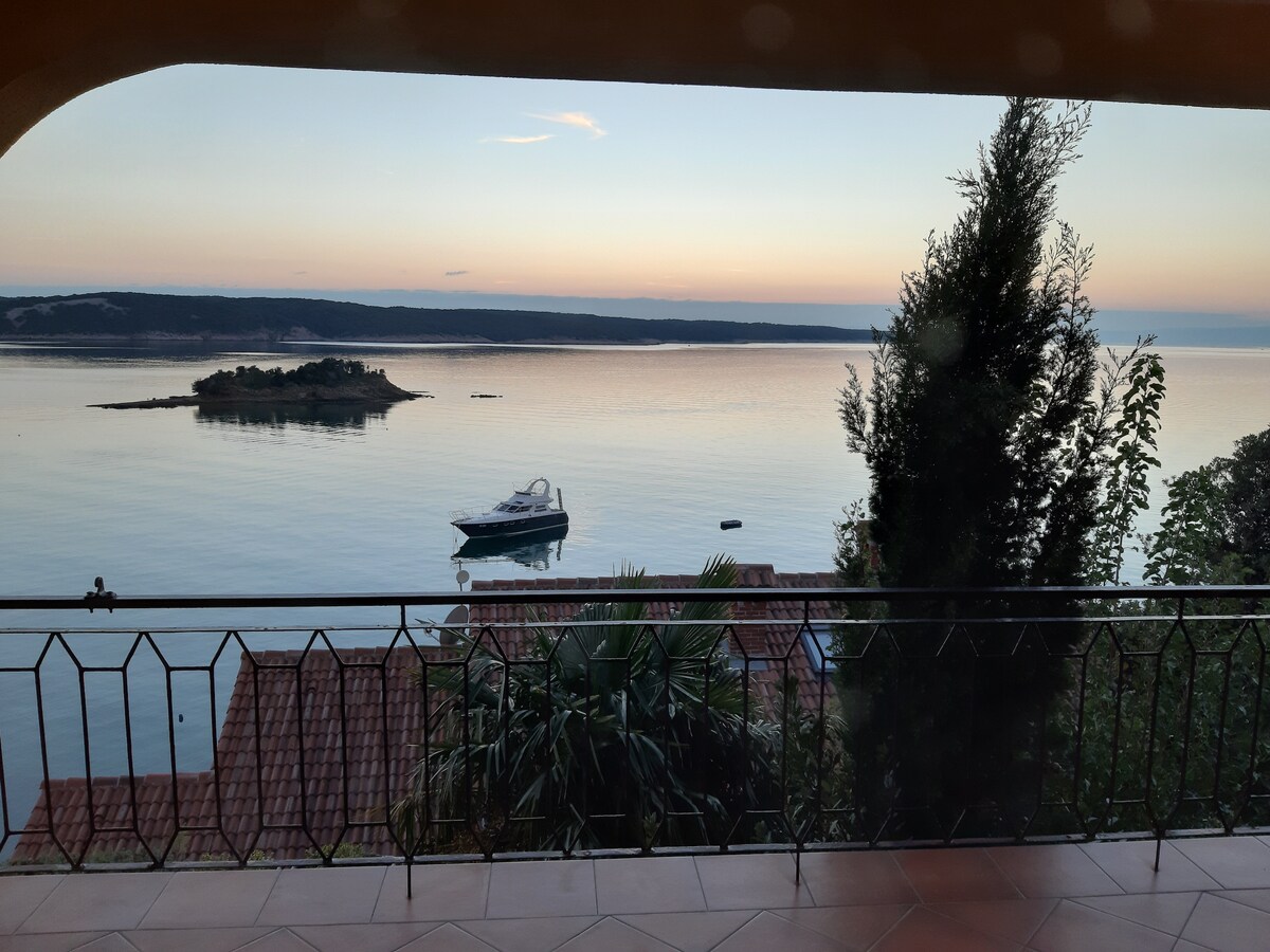 A serene view of the calm sea is captured from a covered balcony. The horizon showcases gentle colors of twilight, while a small island and a boat are visible in the distance. Lush greenery frames the scene, enhancing the coastal ambiance.