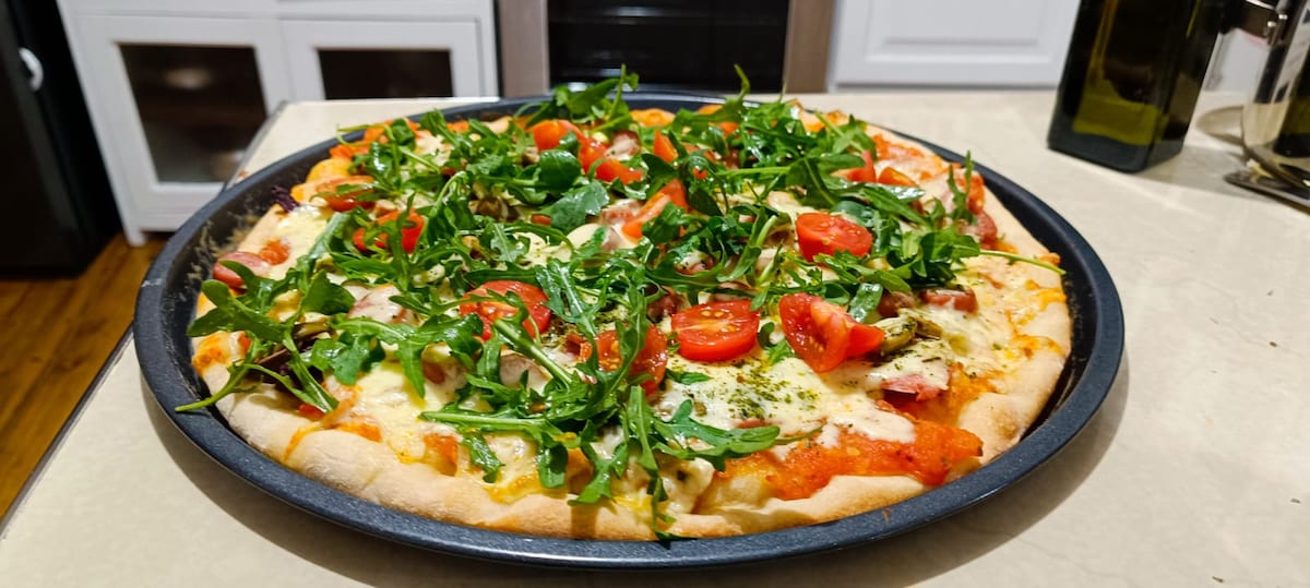 A freshly baked pizza is presented on a round black tray. The pizza is topped with vibrant red cherry tomatoes and a generous layer of green arugula, resting on a bed of melted cheese and tomato sauce. The crust is golden brown and slightly thick.