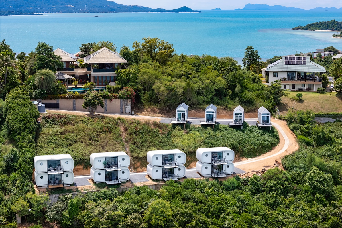 An aerial view captures several tiny houses aligned along a winding path, surrounded by lush greenery. The backdrop features a serene sea and distant hills, while the coastline is visible, emphasizing the property's natural surroundings.