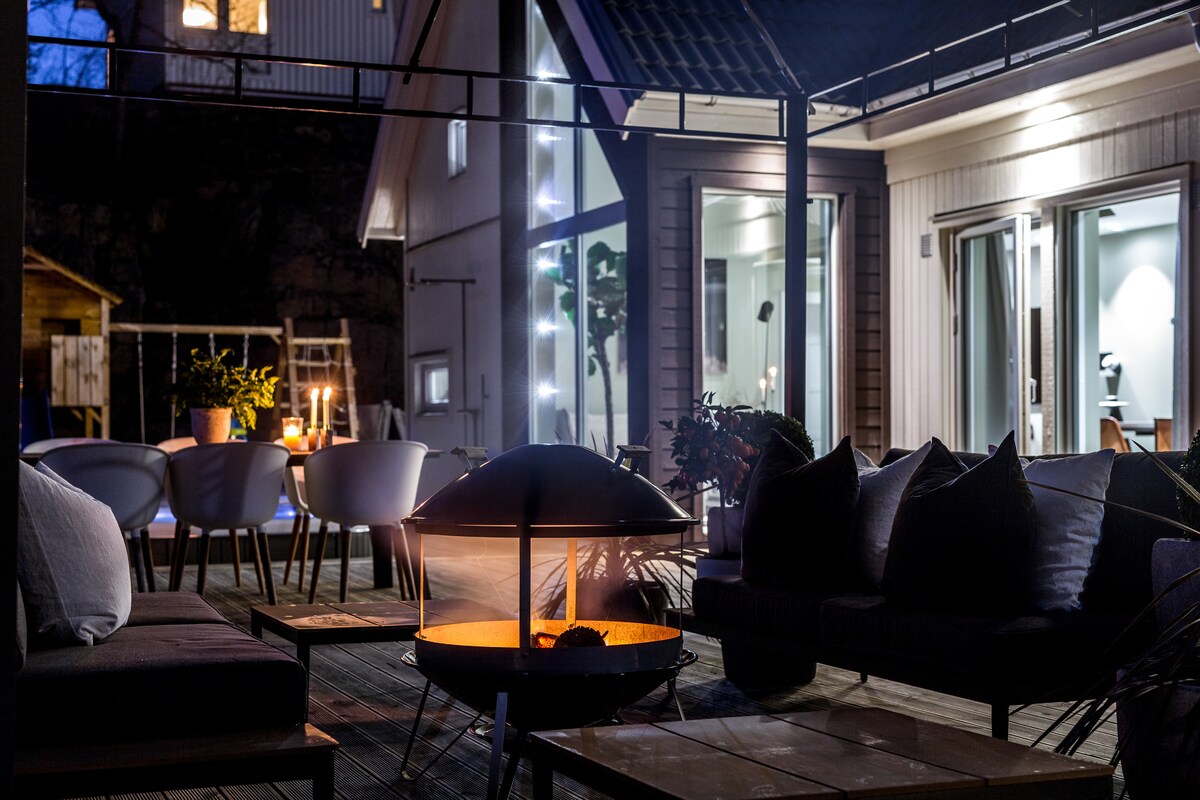 An outdoor seating area features a fire pit as the focal point, surrounded by comfortable lounge chairs and tables. Soft lighting illuminates the space, enhancing the welcoming atmosphere. The home’s interior is visible through large glass doors, blending the indoor and outdoor environments.