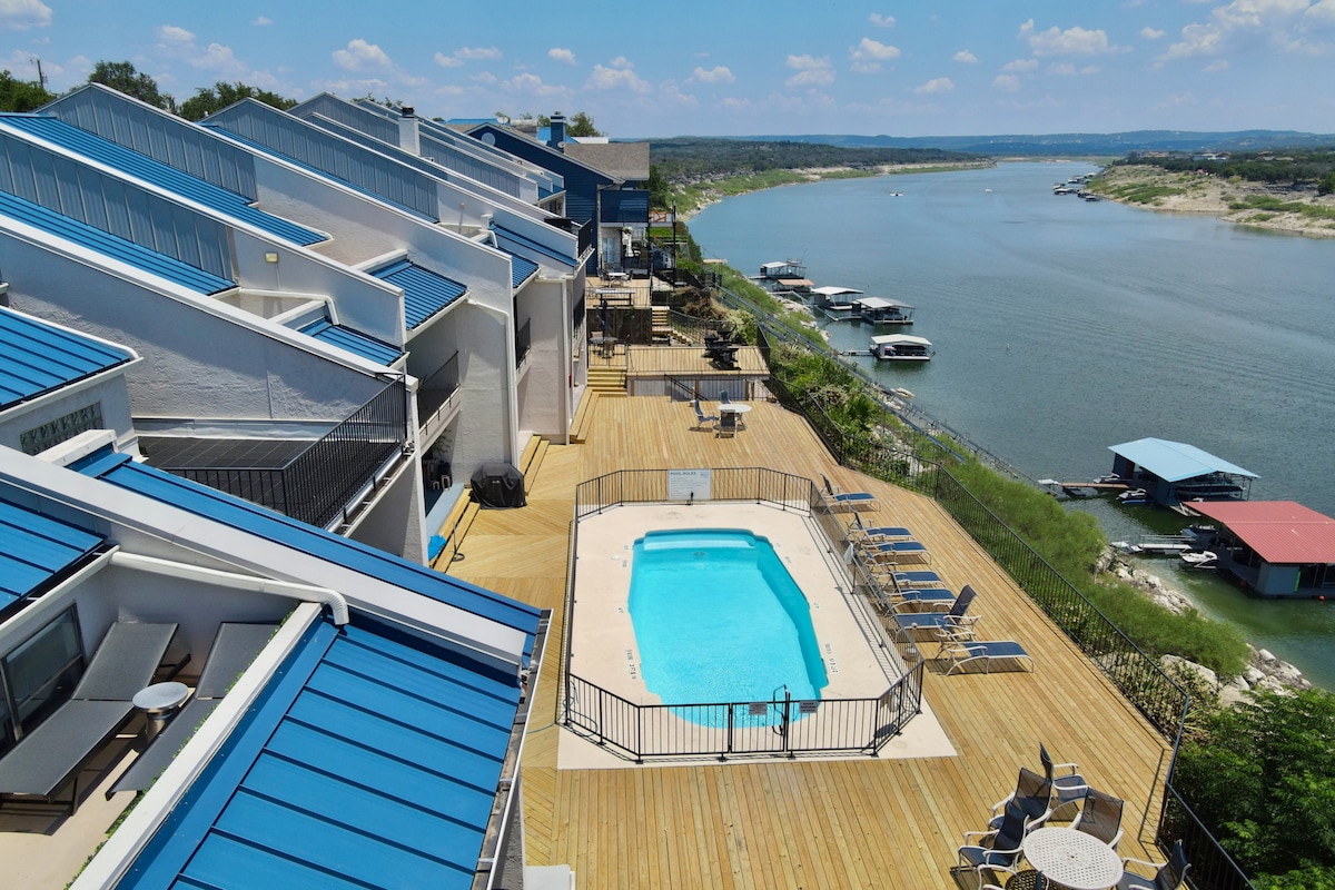Aerial view showcases the inviting pool surrounded by a spacious wooden deck. Loungers are positioned around the pool, while the glistening waters of Lake Travis are visible just beyond. Sunshine fills the scene, with boats gently bobbing in the distance.