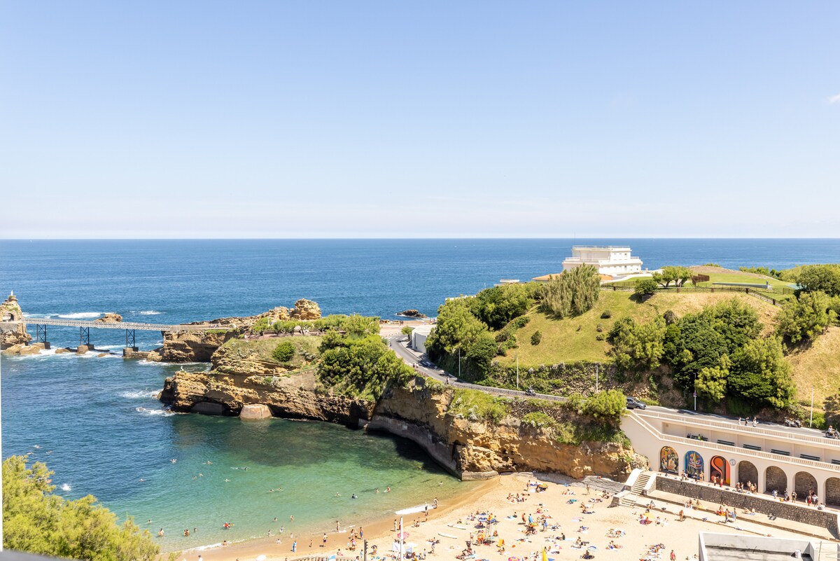 A coastal view captures a sandy beach with people enjoying the sun, surrounded by lush greenery. Waves gently lap at the shore, while distant cliffs lead to the ocean. A clear blue sky complements the tranquil scene, highlighting the natural beauty of this beachside location.