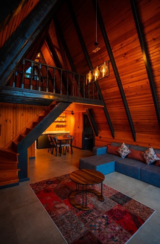 An A-frame living area is presented with wooden walls and beams, creating a warm atmosphere. A spacious corner sofa is complemented by decorative cushions. A round table with four chairs is positioned near the kitchen area, enhanced by pendant lighting hanging from the ceiling.