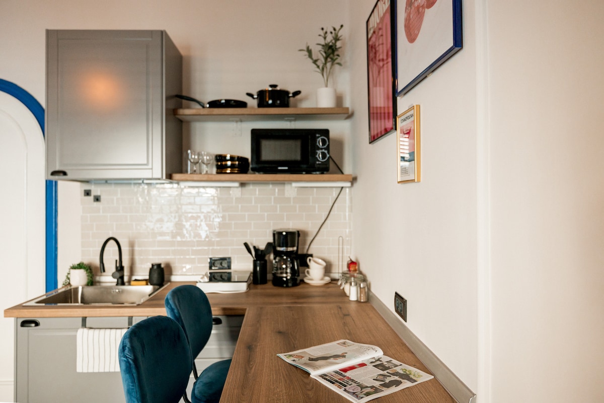 A private kitchenette is showcased with sleek cabinetry and open shelving. Essential appliances including a microwave and coffee maker are neatly arranged. A workspace featuring a wooden countertop and two plush blue chairs is visible, complemented by decorative elements on the wall.