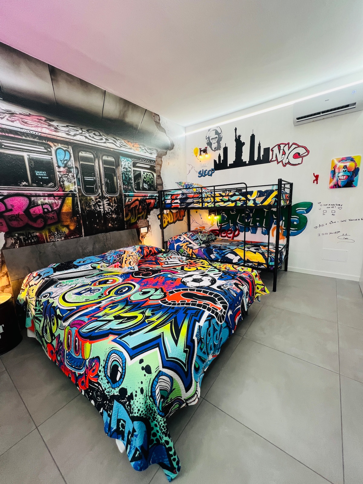 A colorful bedroom is adorned with vibrant graffiti and urban murals inspired by New York City. A spacious bed and a bunk bed are complemented by an artistic bedspread. The décor captures the essence of street art, creating an immersive experience for art enthusiasts.