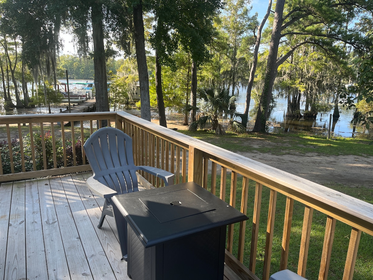 A wooden deck is shown overlooking a serene lakeside view, featuring lush greenery and tall trees. A gray Adirondack chair is placed near a black side table, creating a peaceful seating area to enjoy the natural surroundings.