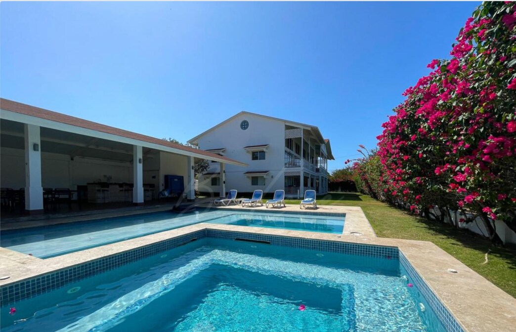 A sunny outdoor area features two swimming pools alongside a white multi-level building. Sun loungers are positioned around the pools, and vibrant bougainvillea plants line the space, adding colorful accents. A covered outdoor dining area is visible, providing shaded seating options.