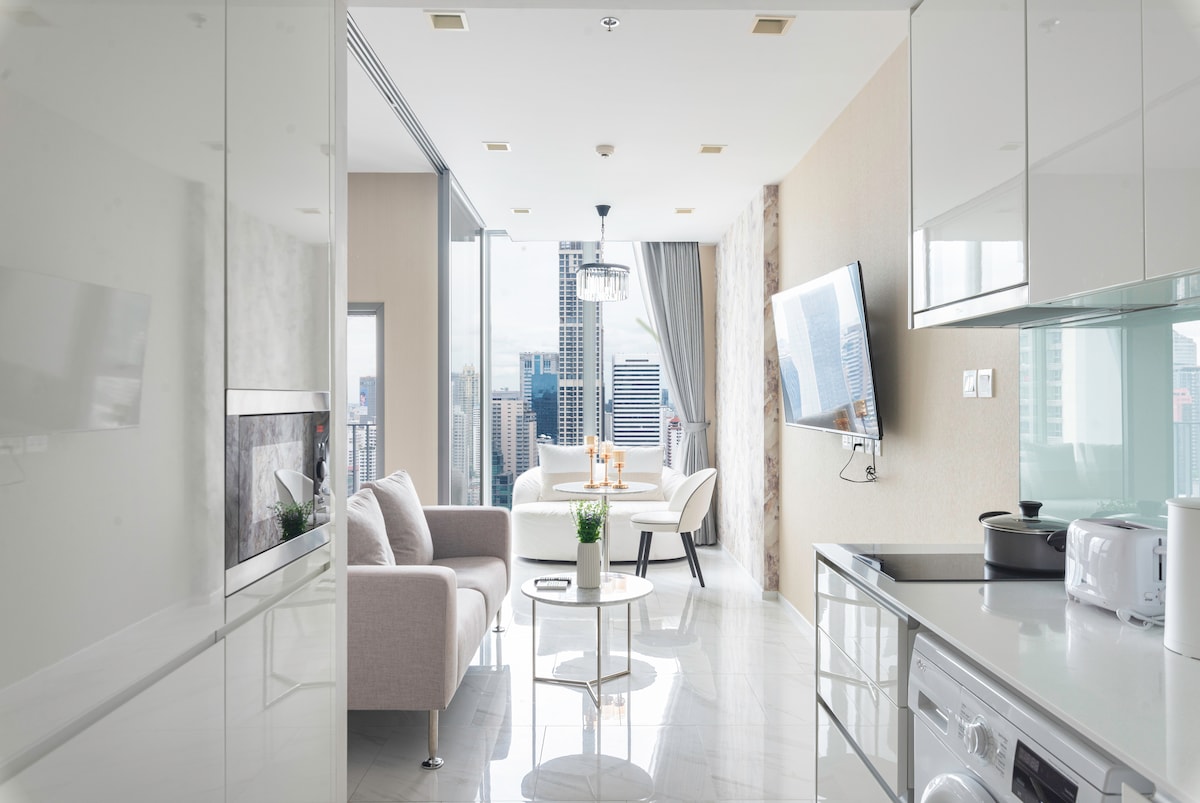 An open living space features a sleek design with white marble flooring. A cozy sofa faces a large flat-screen TV mounted on the wall. Sunlight fills the room through floor-to-ceiling windows, with a dining area showcased in the background, complete with a stylish table and chairs.