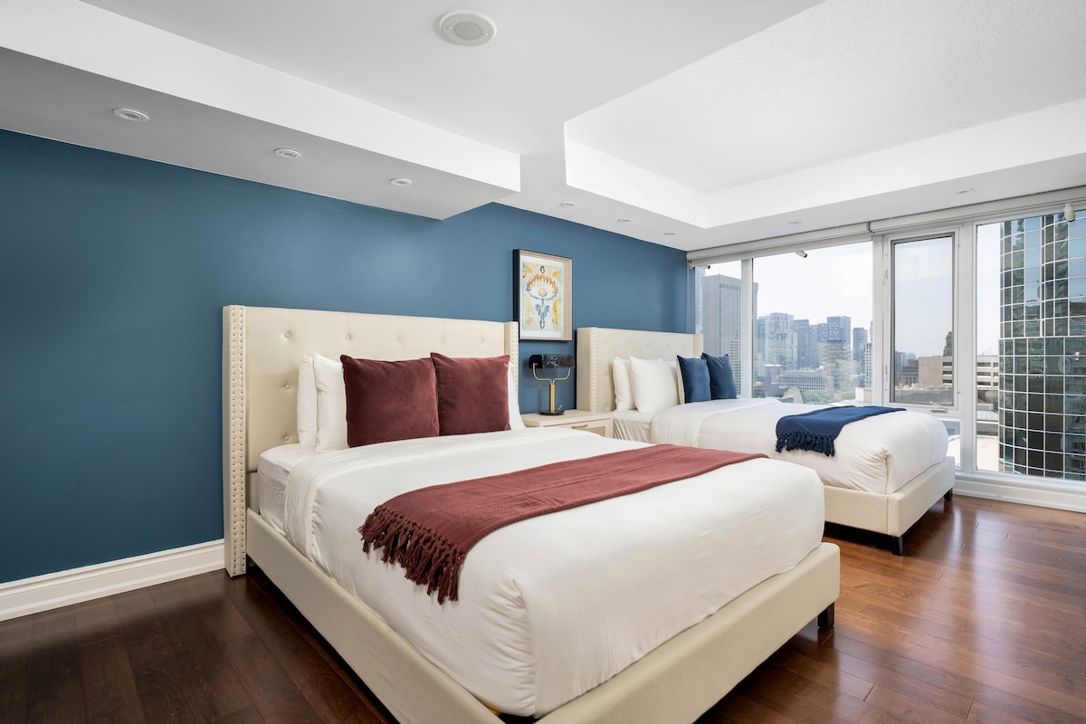 A bright bedroom features two spacious queen beds, each adorned with decorative red and navy throw blankets. A large window offers sweeping views of the city skyline, while a contemporary art piece is displayed on the blue accent wall.