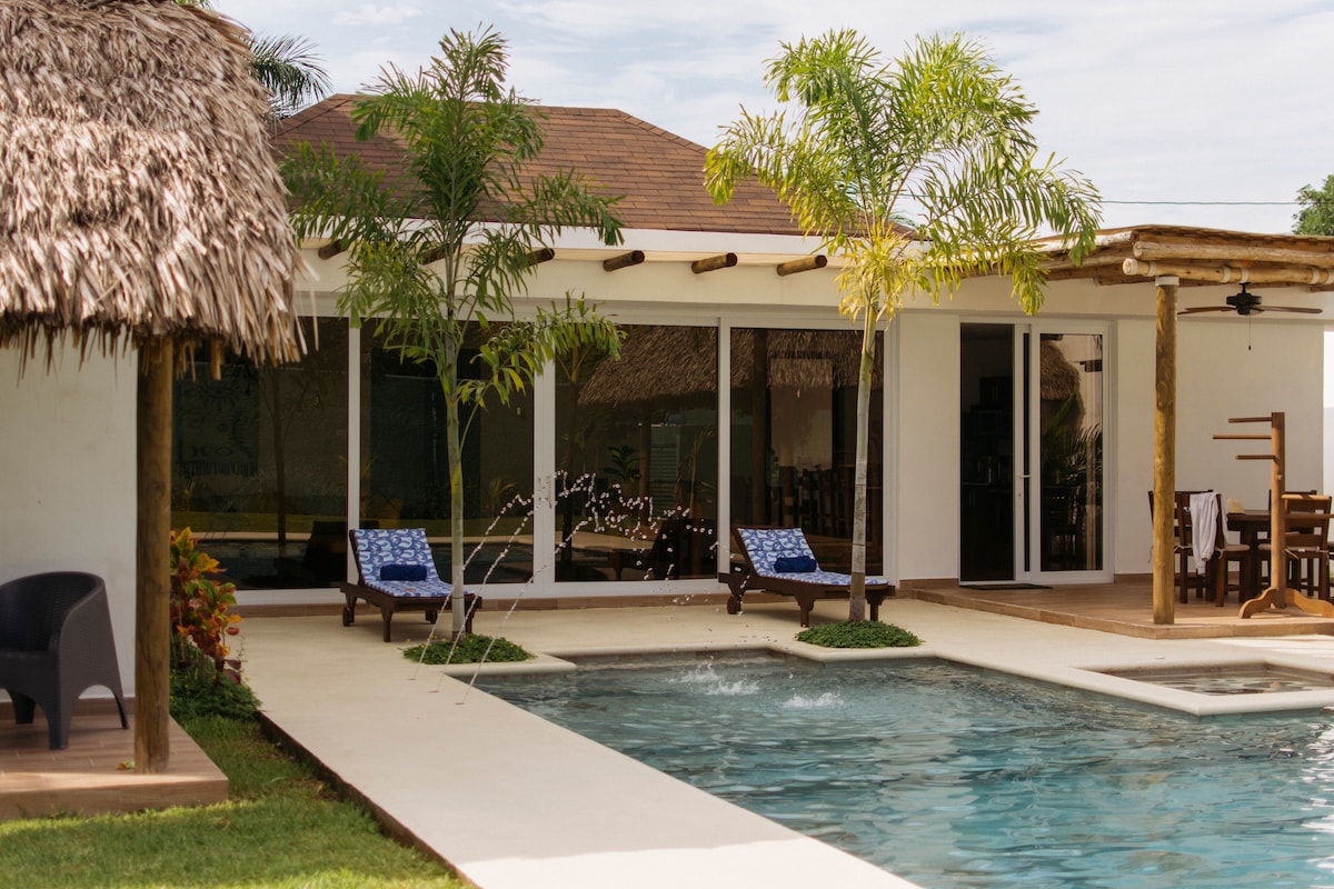 An outdoor area features a shimmering pool surrounded by lush grass and tropical plants. Two blue lounge chairs are positioned beside the water, while a thatched-roof structure sits nearby. Large glass doors open to a spacious indoor area, enhancing natural light.