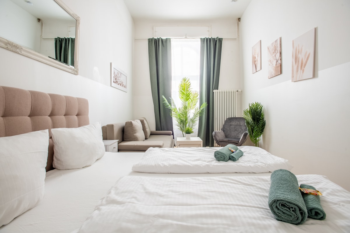A bright and inviting bedroom is showcased with a spacious bed dressed in soft linens. Two decorative plants add a touch of greenery, while a comfortable seating area features a light-toned couch and chair. Natural light filters through the large window, complemented by flowing curtains.