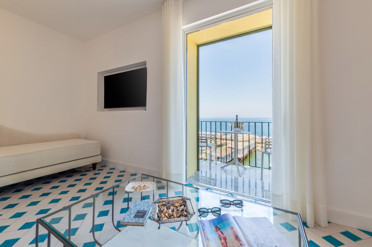 The living area is highlighted by a large glass coffee table displaying assorted snacks and magazines. A comfortable sofa faces a wall-mounted television. Natural light filters through sheer curtains, illuminating the space and revealing a view of the sea through an open door.