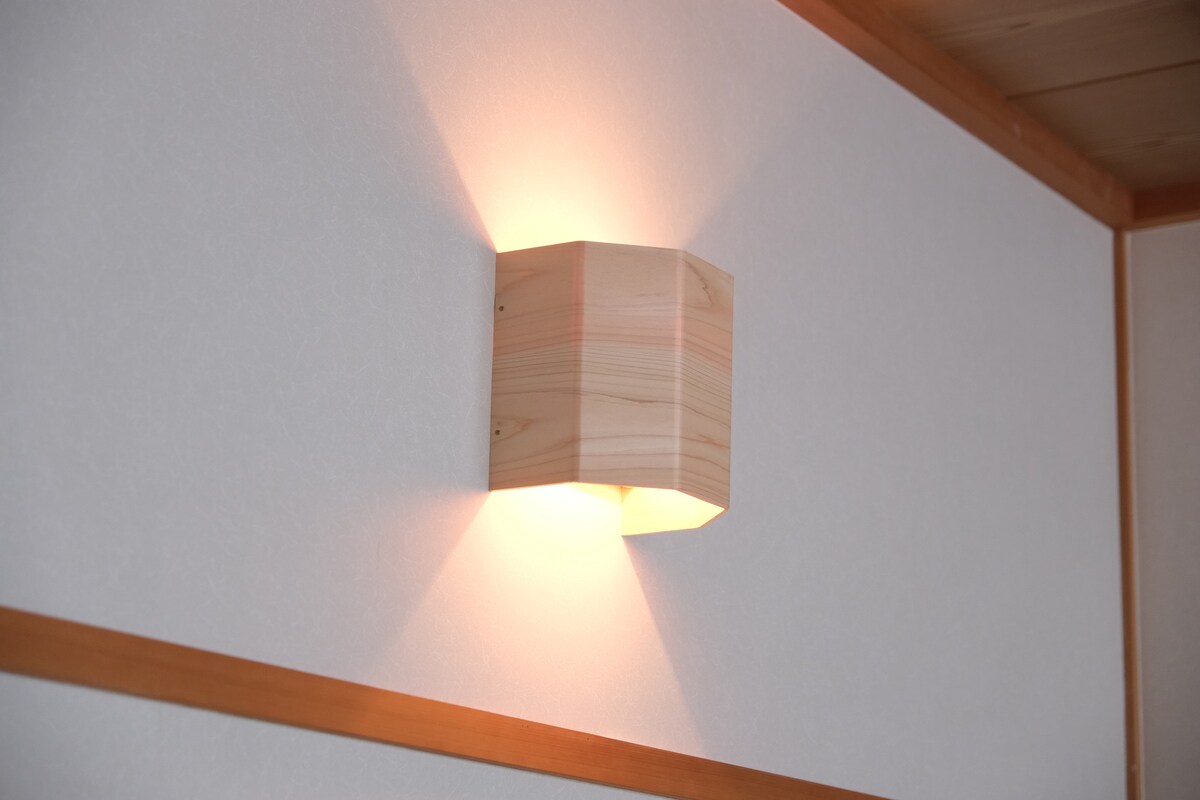 A warm wooden sconce is mounted on the light-colored wall, casting a soft, ambient glow. The design features a rectangular shape with rounded edges, enhancing the simplicity of the space while providing gentle illumination.