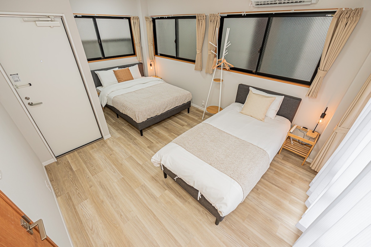 A well-lit bedroom features two beds, one spacious and one single, positioned against light-colored walls. Natural light enters through large windows, adorned with soft curtains. A minimalist chair and bedside table complete the serene atmosphere, providing a cozy space for relaxation.
