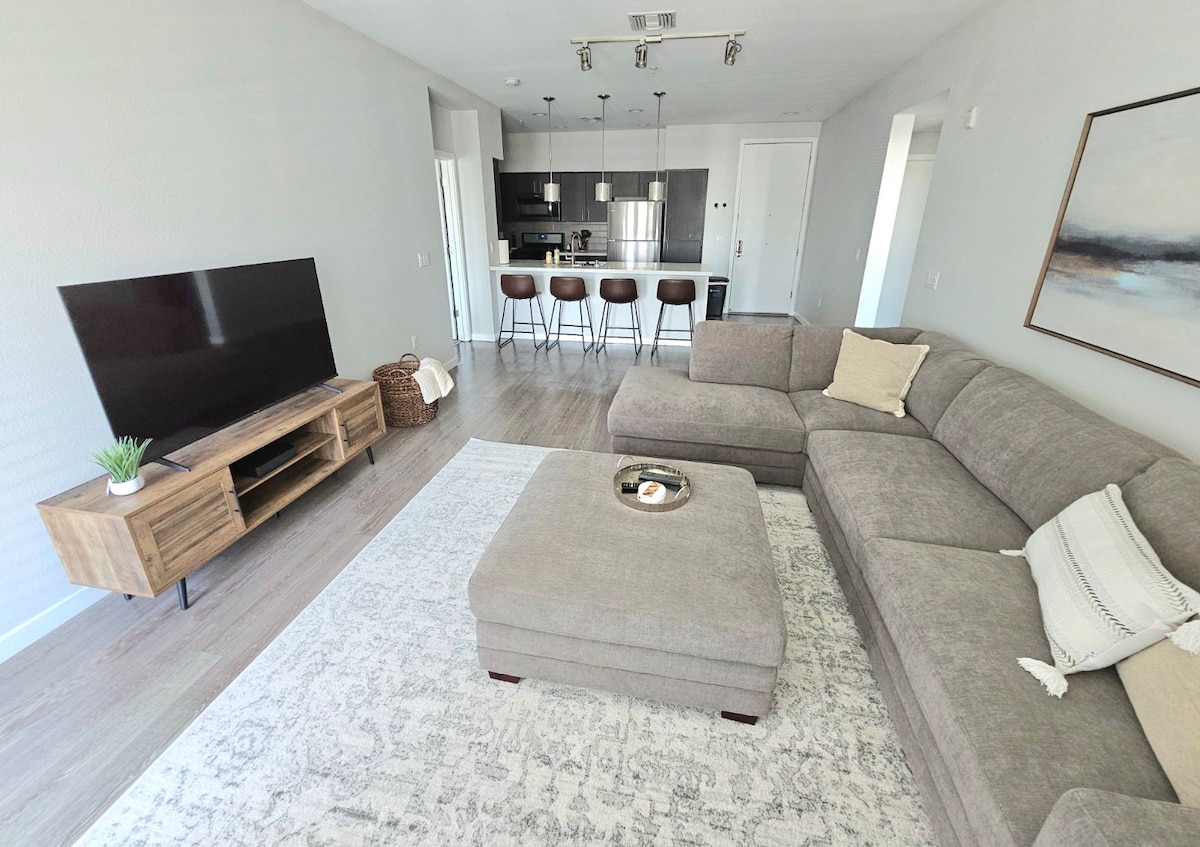 A modern living area features a comfortable sectional sofa and a coffee table with decorative items. A flat-screen television is mounted on a wooden media console. The open layout leads to a sleek kitchen with bar seating for four, enhancing the spacious feel of the room.