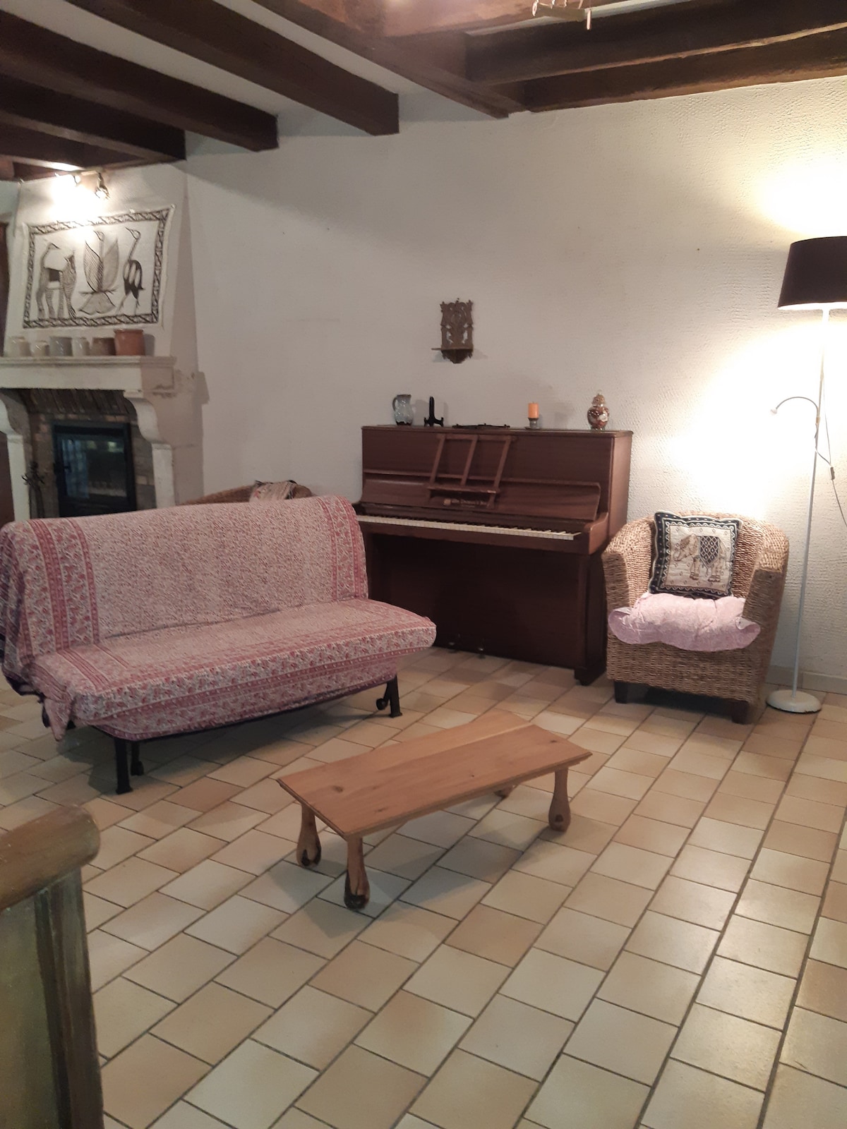An inviting living area features a patterned sofa and a cozy armchair. A wooden coffee table sits centrally, complemented by a piano against the wall. Warm lighting is provided by a floor lamp, while textured tiles add a homely touch to the space.