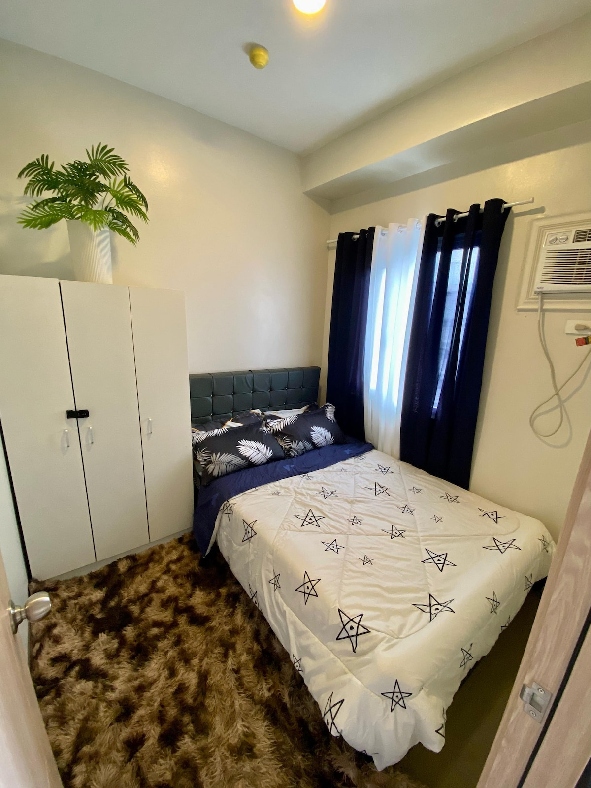 A cozy bedroom features a comfortable bed with patterned bedding. A small wardrobe stands against the wall. Soft, plush carpeting covers the floor, and a potted plant adds a touch of greenery. Natural light filters through dark curtains, creating a serene ambiance.