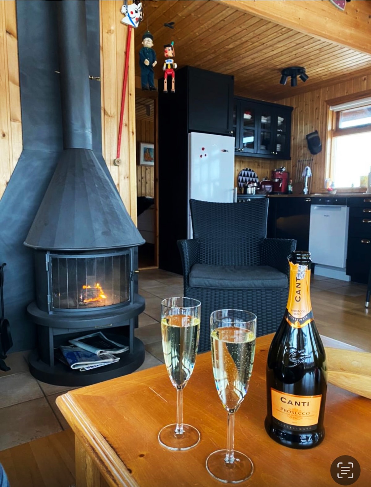 A wooden interior features a cozy sitting area with a glass coffee table, two flutes filled with sparkling wine, and a bottle nearby. A modern fireplace with a black chimney provides warmth, complemented by woven black chairs. The kitchen area is visible in the background.