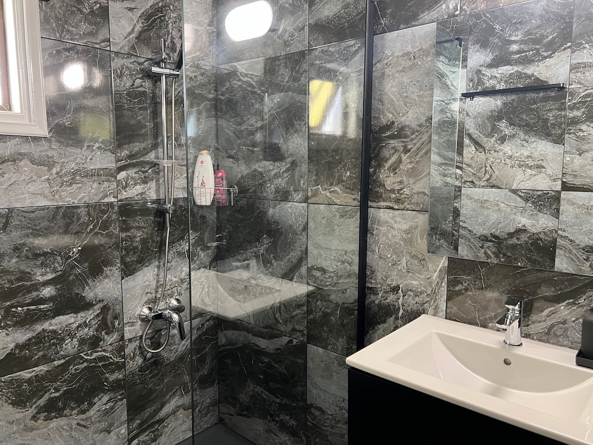 A modern bathroom features sleek grey marble walls and a glass-enclosed shower. The shower is equipped with a handheld and wall-mounted faucet. A well-lit mirror is situated above a minimalist sink with a dark cabinet below, providing ample space for toiletries.