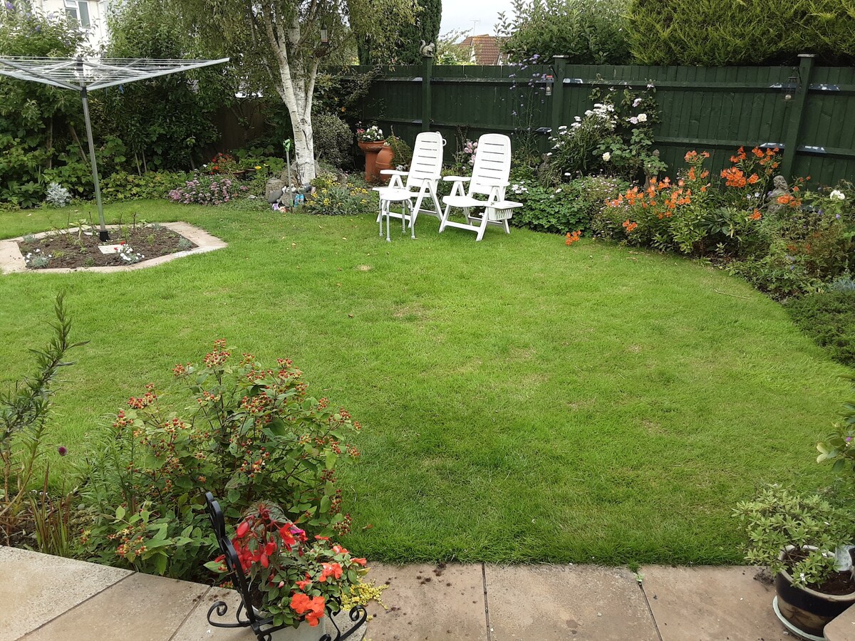 A well-maintained garden displays lush green grass surrounded by colorful flower beds. Two white lounge chairs are positioned on the lawn, providing a cozy spot for relaxation. A patio area with stone tiles leads to the garden, enhancing the outdoor space.