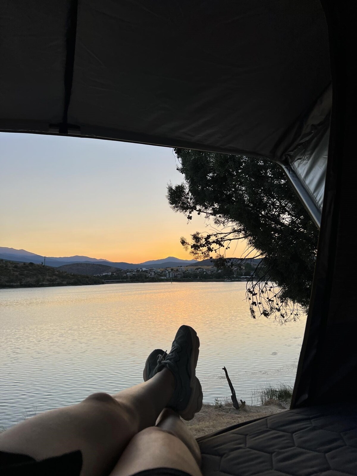The interior of a tent frames a serene view of a calm lake at sunset. Silhouetted mountains rise in the distance, while the soft colors of twilight reflect on the water. A pair of feet in sneakers can be seen resting in the foreground.