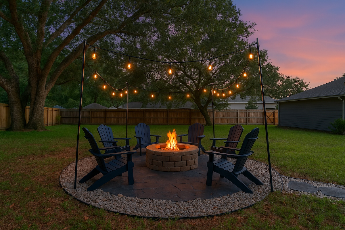 A cozy firepit area is surrounded by four Adirondack chairs, set on a stone patio. Soft string lights are hung overhead, casting a gentle glow in the evening twilight. Lush green grass and trees create a serene backdrop for outdoor gatherings.