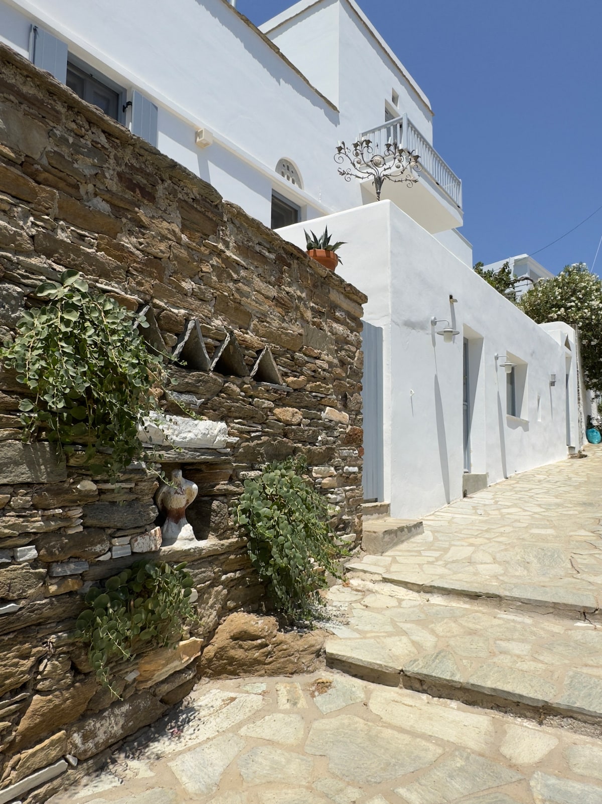 A narrow stone pathway leads to a whitewashed building. The rustic stone wall features vertical planters with greenery. Sunlight casts natural shadows, highlighting the texture of the stones and the simplicity of the architecture.