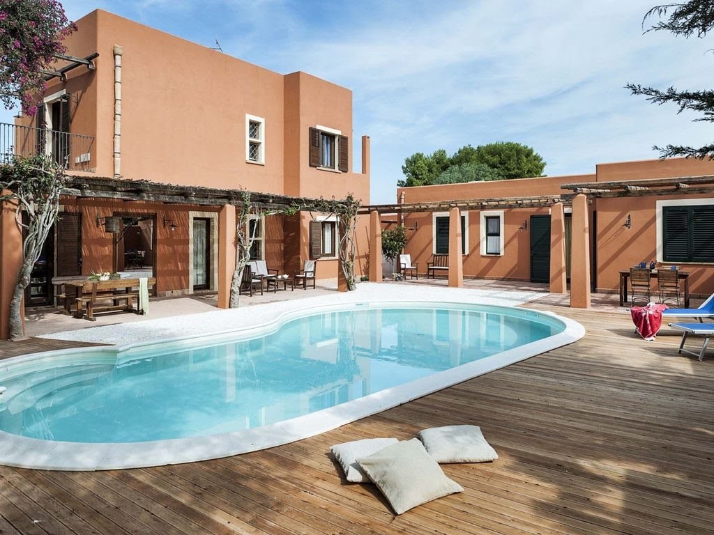 A private outdoor area features a serene swimming pool surrounded by wooden decking. Sun loungers and floor cushions are arranged nearby, inviting relaxation. The villa's warm-toned exterior complements the natural surroundings, with moments of shade provided by nearby trees and structures.