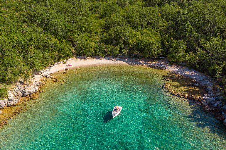 A serene view of a secluded pebble beach bordered by lush greenery. The clear turquoise water enhances the calm atmosphere, while a small boat is anchored nearby, offering a glimpse of tranquil outdoor exploration.