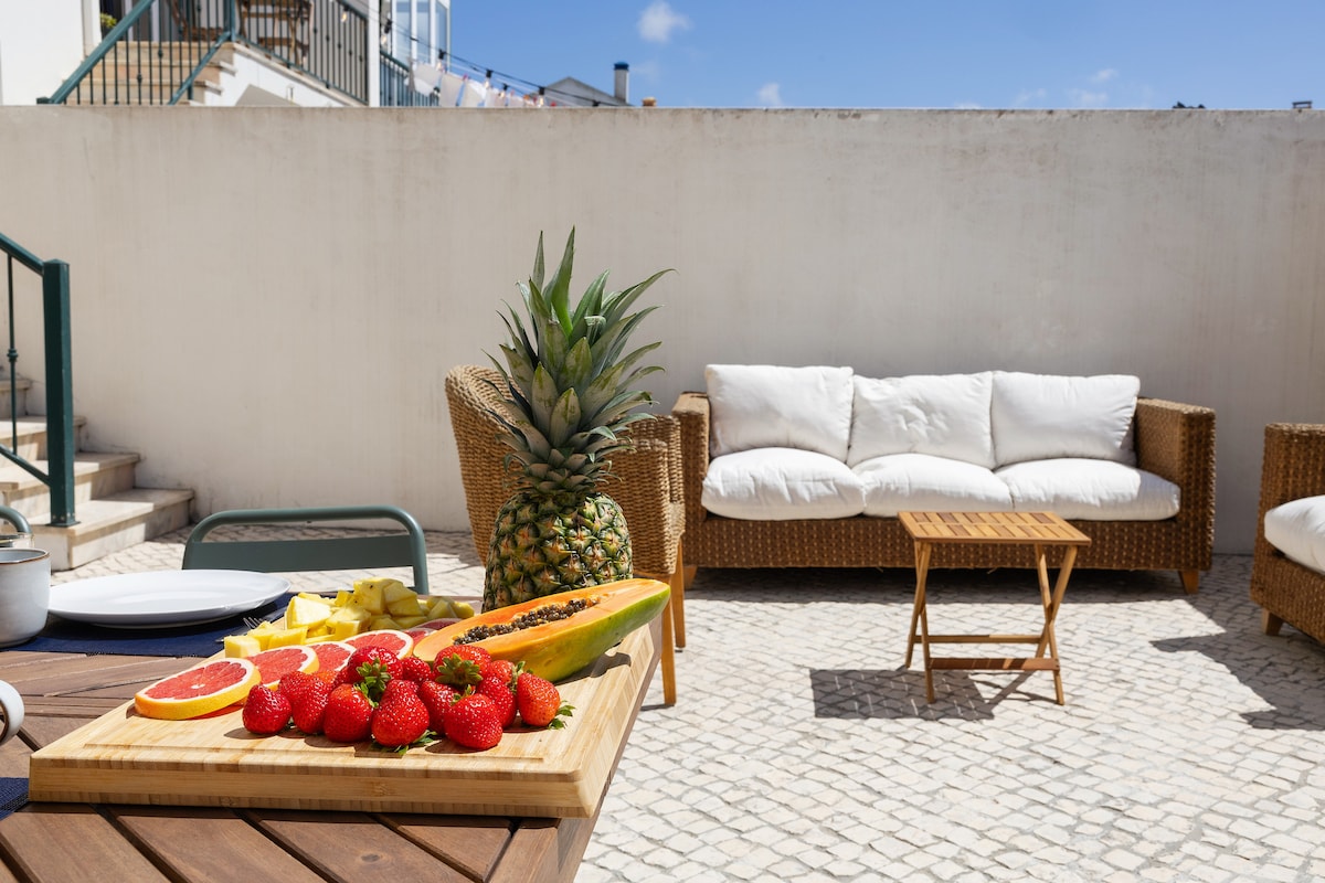 An outdoor terrace features comfortable wicker seating accompanied by a small wooden table. A selection of tropical fruits is artfully arranged on a wooden cutting board. Sunlight illuminates the space, enhancing the inviting and peaceful atmosphere.