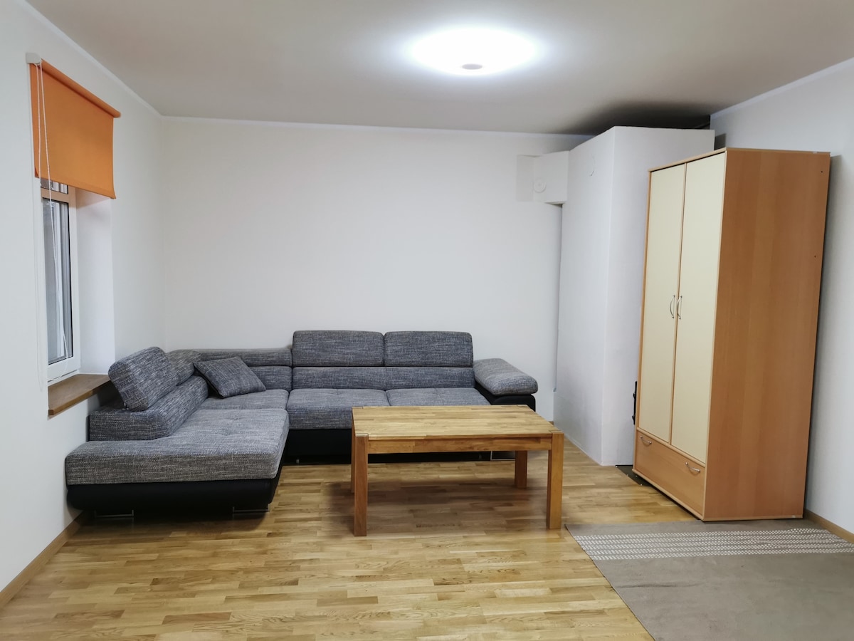 A spacious living area features a modern, grey fold-out sofa complemented by a wooden coffee table. A light orange window shade adds a touch of color, while a cream-colored wardrobe stands against the wall, creating a functional and simple environment.