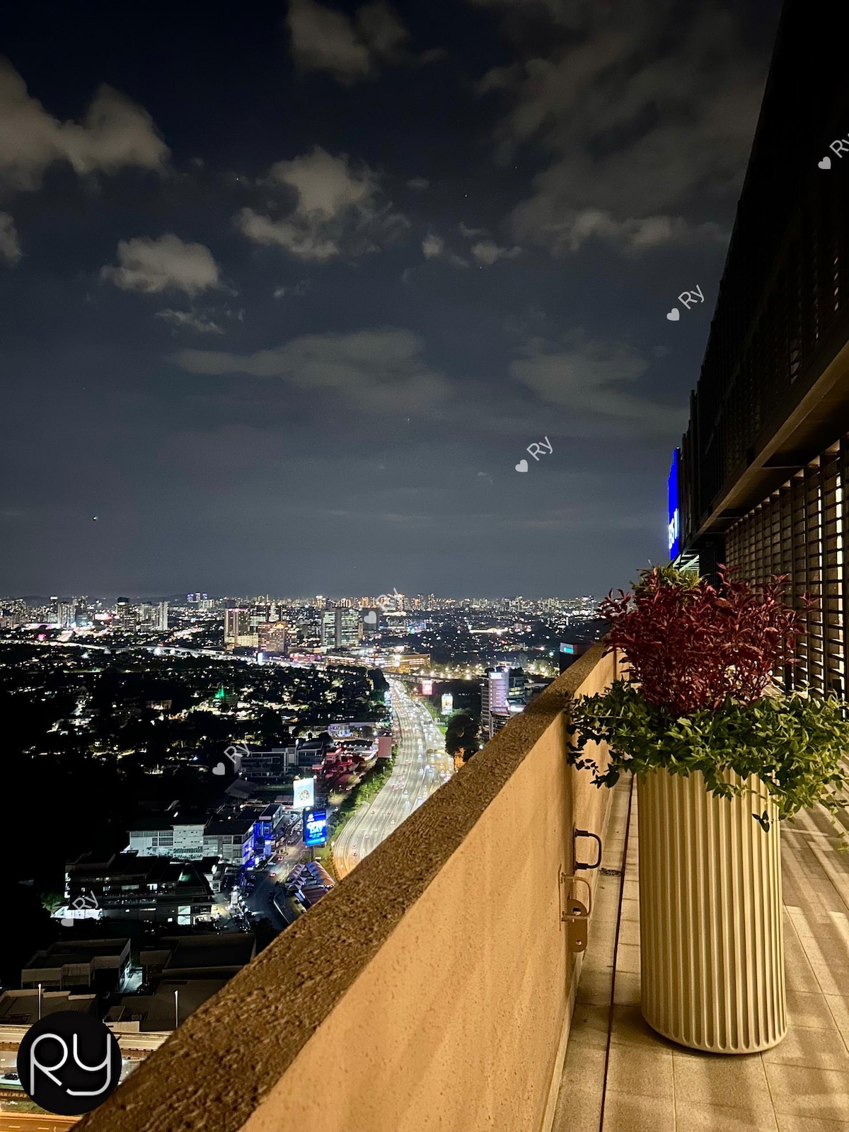 A city view is captured from a high balcony, revealing a sprawling landscape of illuminated buildings and streets below. Potted greenery adds a touch of color to the sleek outdoor area, with the night sky stretching overhead and soft clouds drifting in the distance.