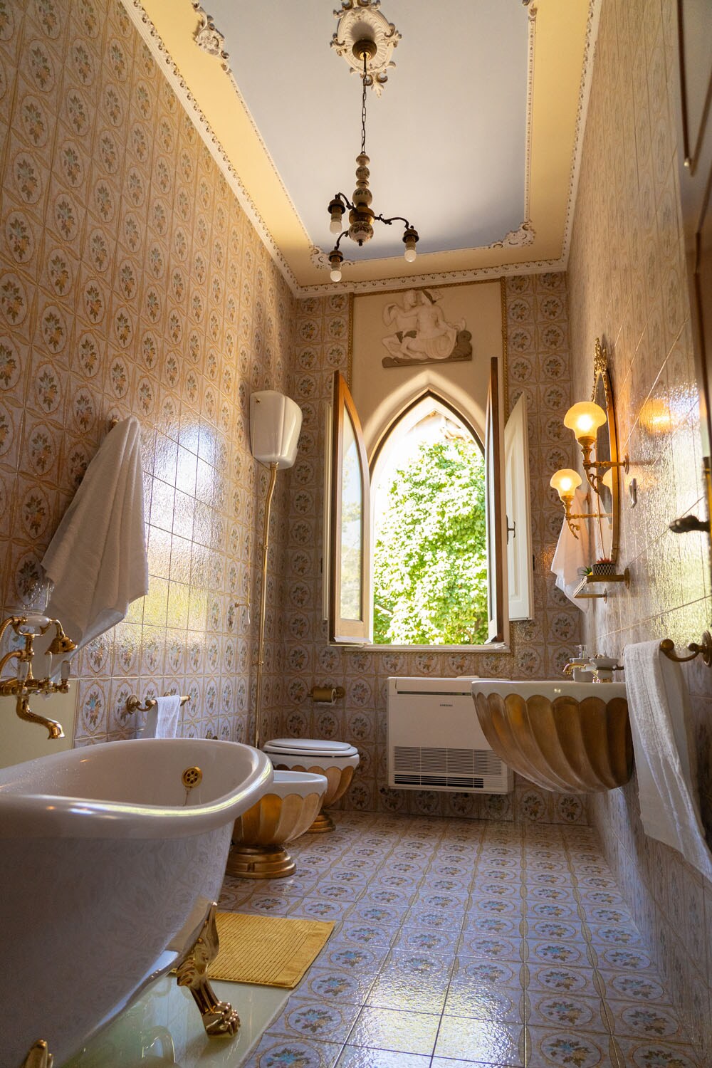 The elegant bathroom features ornate tiles and a vintage bathtub, complemented by a decorative sink. Natural light filters through an arched window, providing a view of greenery outside. Classic light fixtures enhance the room's historical charm.