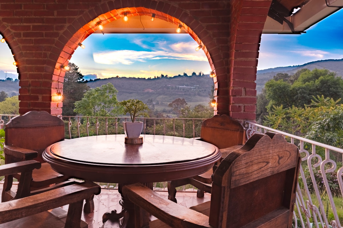 An inviting terrace features a round table surrounded by four wooden chairs, with a small potted plant at the center. The arched brick structure provides a cozy frame, while views of rolling hills and the sky add a serene backdrop.