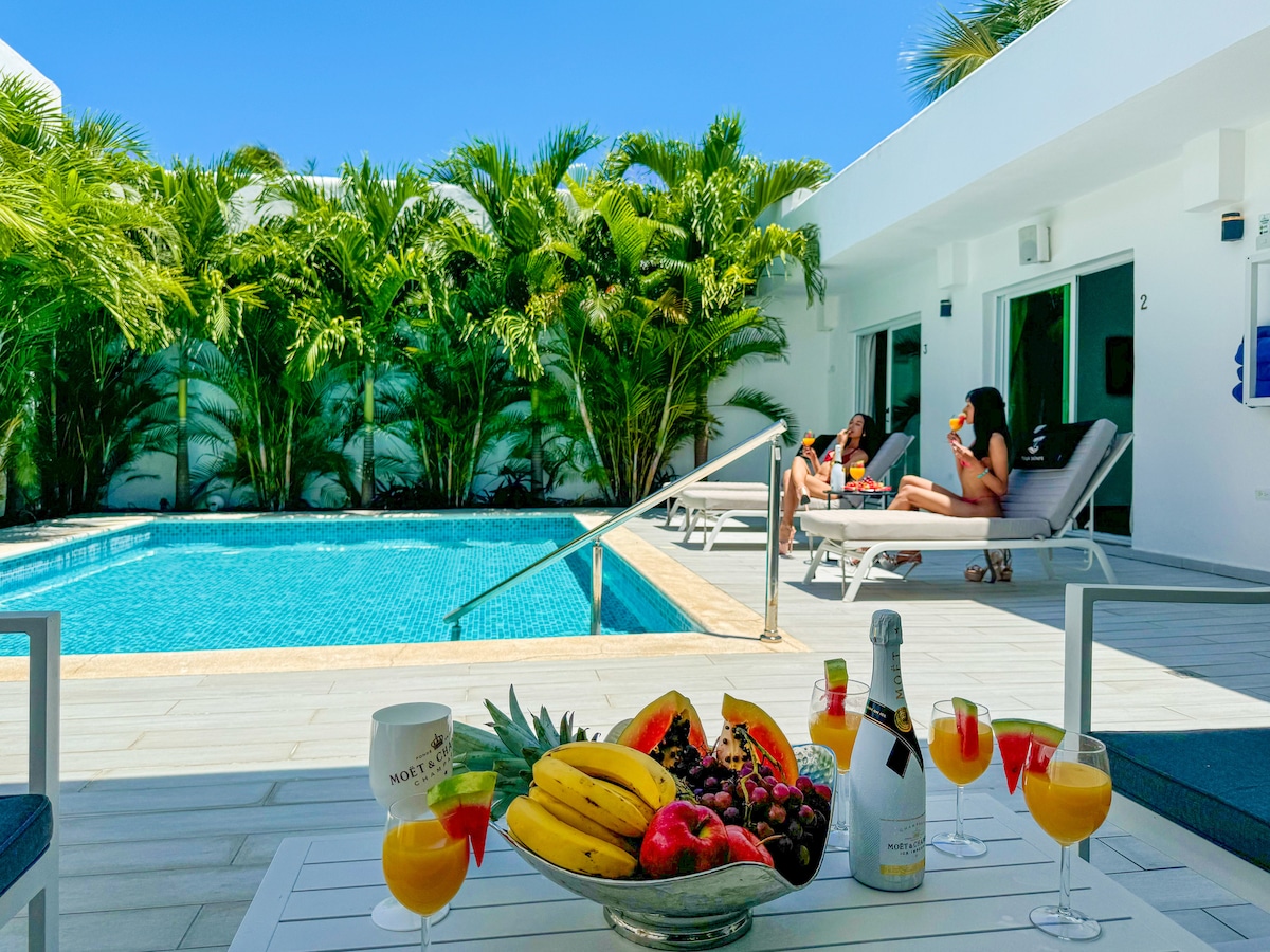 A refreshing private pool area is surrounded by lush tropical plants. Two individuals lounge on comfortable sunbeds, enjoying the sunshine. A table with vibrant cocktails and a fruit platter is placed in the foreground, inviting relaxation and leisure.