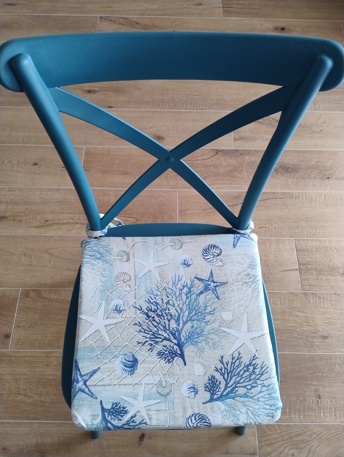 A wooden chair with a cross-back design features a cushion adorned with marine-themed patterns, including blue corals and seashells. The chair is positioned on a light wooden floor, highlighting the calm and nature-inspired aesthetic of the space.