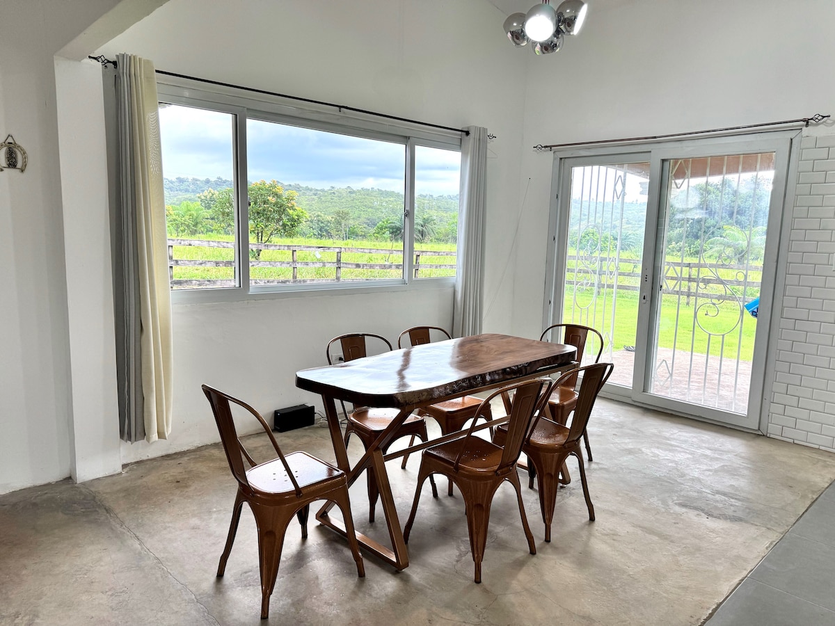 A dining area is featured with a long wooden table accompanied by six wooden chairs. Large windows allow natural light to fill the space, offering views of the surrounding greenery. A sliding door leads to an outdoor area, enhancing the connection with the natural environment.