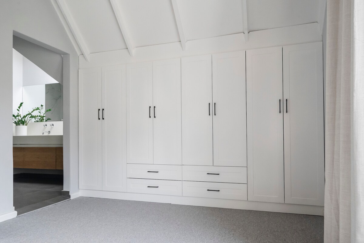 A minimalistic and bright bedroom features a wall of white cabinetry with sleek black handles, providing ample storage. The light-colored carpet complements the room’s airy feel. A doorway to the left leads to a modern bathroom area, enhancing the overall spaciousness.