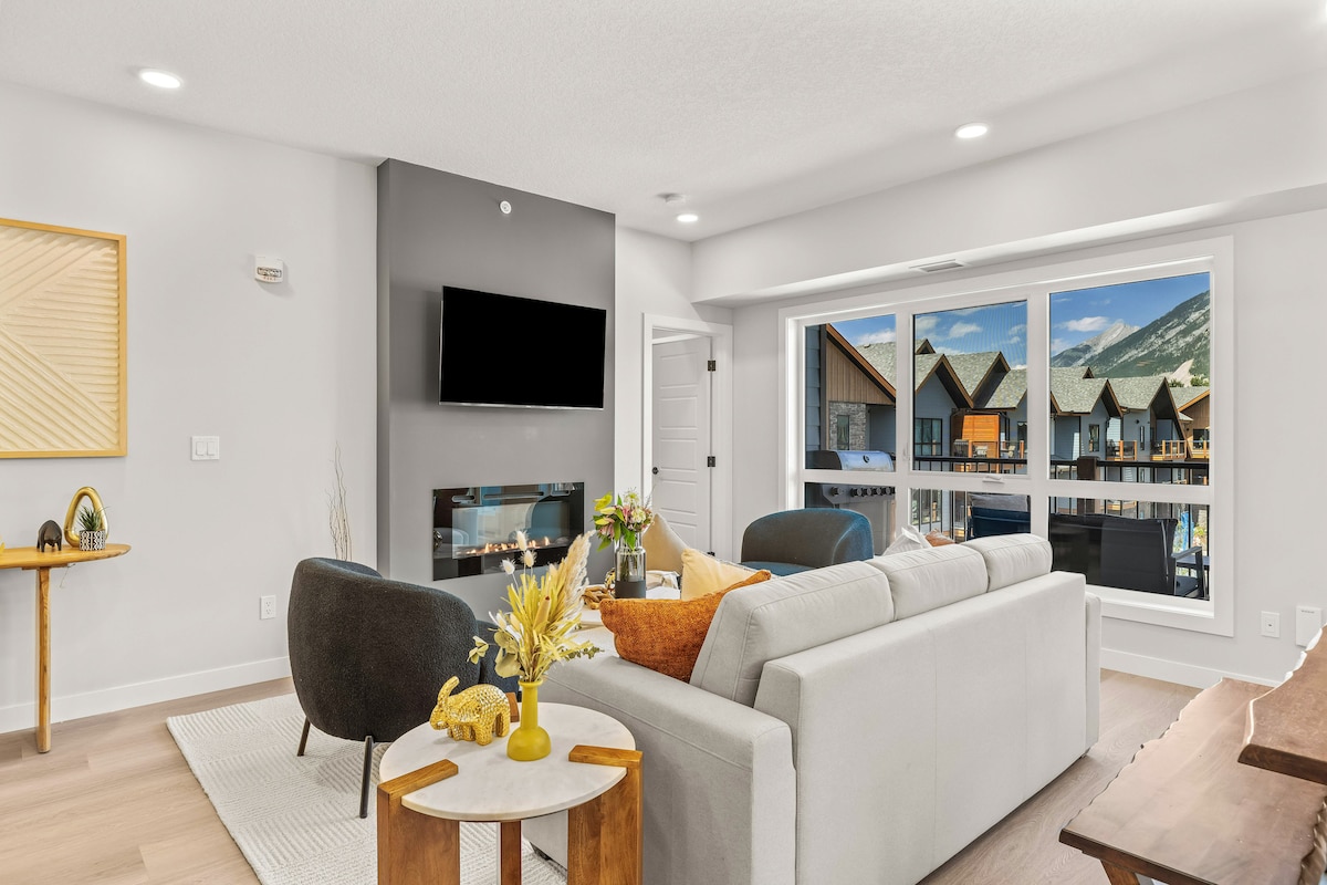 The living area showcases a spacious sofa, accent chairs, and a modern fireplace. A wall-mounted TV is visible, while large windows offer scenic mountain views. Natural light fills the space, creating a comfortable atmosphere for relaxation and gatherings.