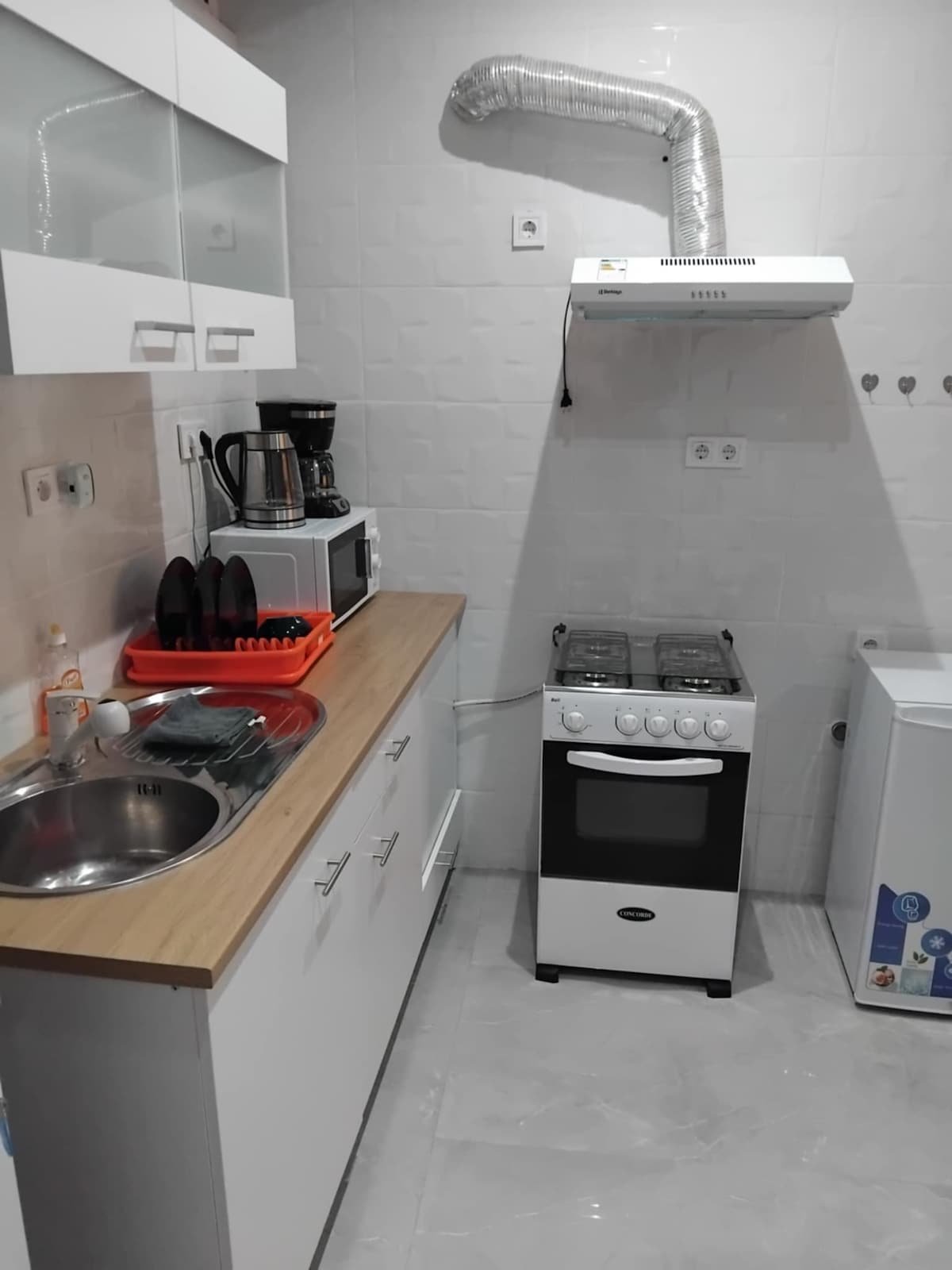 A compact kitchen is equipped with essential appliances, including a stovetop oven, coffee maker, and microwave. A stainless steel sink is positioned under a wall cabinet, while a dish rack holds plates. Light-colored cabinetry and a wooden countertop create a functional cooking space.