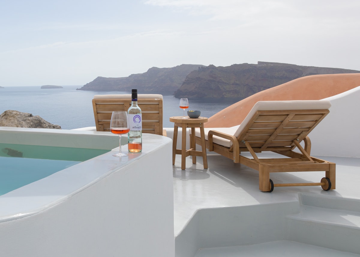 Two wooden lounge chairs are positioned beside a plunge pool, with a small table holding a wine glass and a bottle of wine. The view overlooks the Aegean Sea and distant volcanic islands, with soft pastel hues accenting the landscape.