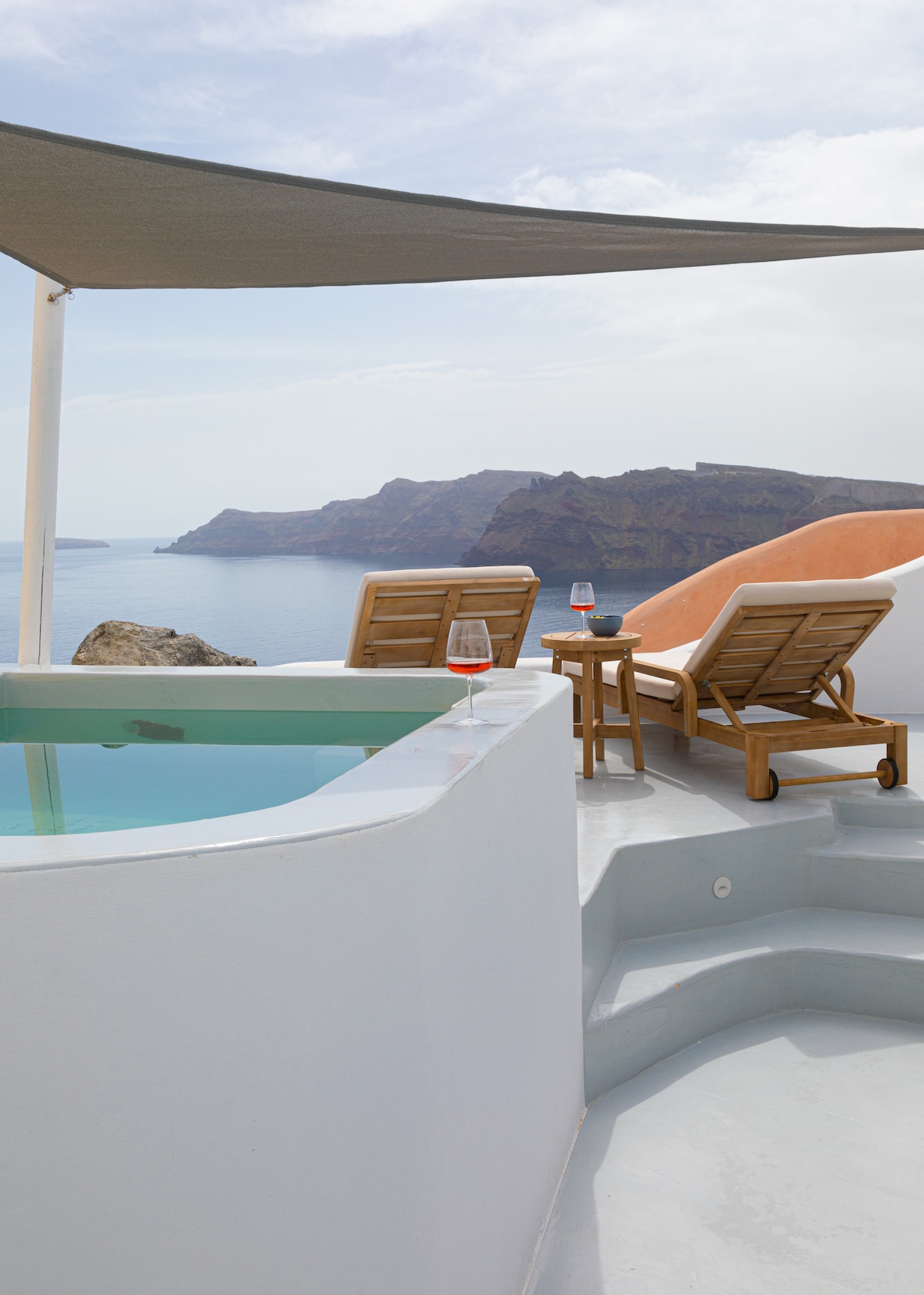 A private plunge pool is positioned at the cliff's edge, offering views of the Aegean Sea. Two wooden lounge chairs are placed nearby, accompanied by a small table with glasses. A shaded area is provided by a canopy, enhancing the scenic atmosphere.