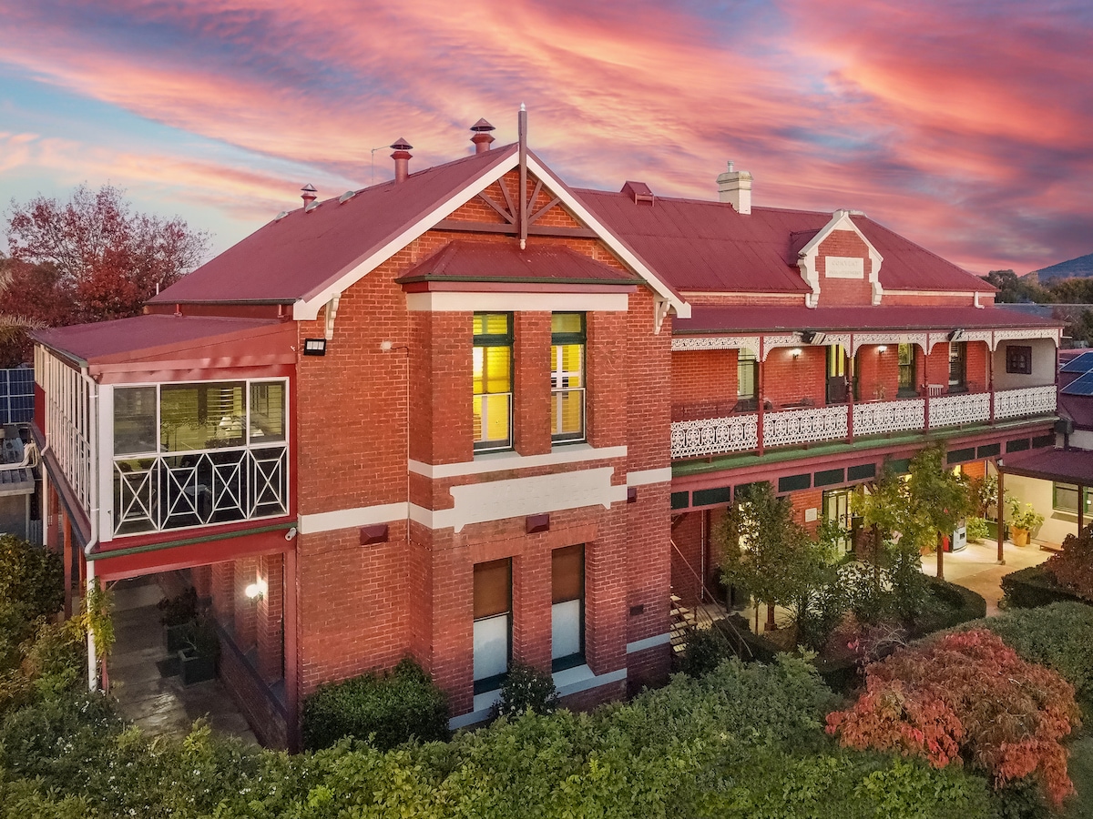The red brick exterior of the resort features a vintage architectural style, complemented by a deep red roof and decorative balcony railings. Lush greenery surrounds the building, and the sky is painted in warm hues during sunset, creating a serene backdrop.